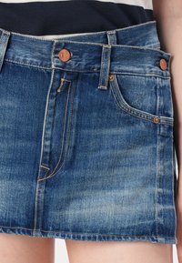 Jeanskjørt i mellomblå vask med en falmet overflate. Har fem lommer, kontrasterende sømmer og metallknapplukking i livet.