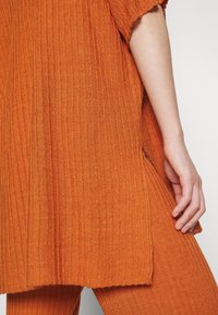 Kädet lepäävät oranssin poimuttuneen neulepaidan, jossa on sivuhalkio ja siihen sopivat poimuttuneet housut, päällä tasaisella taustalla.