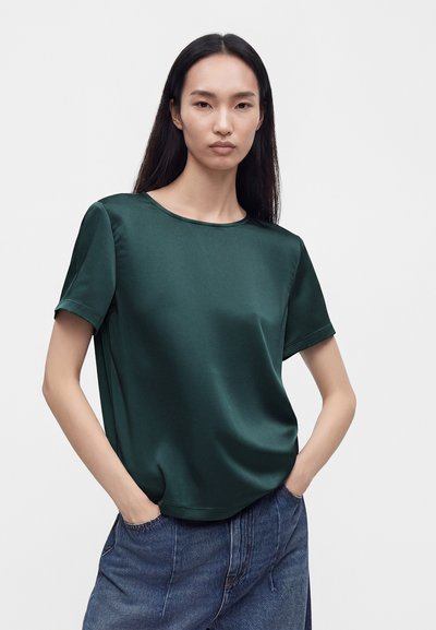 Femme aux longs cheveux foncés portant un haut court en satin vert foncé à manches courtes et un jean bleu, debout les mains dans les poches devant un fond blanc.