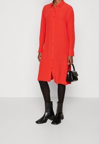 Abito camicia rosso con maniche lunghe, colletto e taglio dritto. Abbinato a collant neri e stivali con tacco spesso. Piccola borsa a tracolla nera.