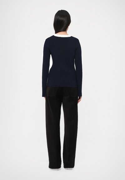 Suéter azul marino con textura acanalada, mangas largas y cuello blanco; combinado con pantalones de pana negros. Textura lisa y diseño ajustado.