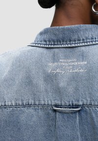 Giubbotto di jeans azzurro chiaro con colletto, cuciture visibili e un'etichetta intrecciata con testo bianco. Design classico e tessuto testurizzato.