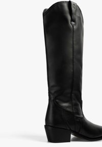 Bota de cuero negra hasta la rodilla con punta afilada, tacón bajo y detalle de costura vertical a lo largo del lado. Textura suave con un acabado pulido.