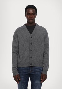 Grauer Cardigan mit Schalkragen, gerippten Säumen und Bündchen, mit sechs schwarzen Knöpfen auf der Vorderseite. Getragen über einem schwarzen Hemd.