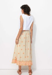 Jupe maxi légère, beige avec des motifs géométriques orange, ornée d'un ourlet décoratif et d'une taille élastique. Associée à un haut blanc.
