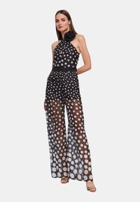 Jumpsuit nero a pois con tessuto trasparente, collo all'americana e grande fiore decorativo. Presenta gambe larghe e fluide con pois bianchi a contrasto.