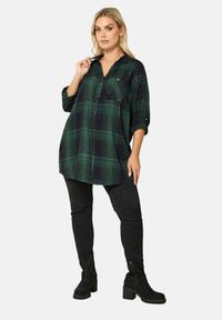 Zielona i granatowa koszula tunika w kratę z dekoltem w serek, podwiniętymi rękawami i przednią kieszenią. Nosi się ją z czarnymi spodniami i botkami za kostkę.