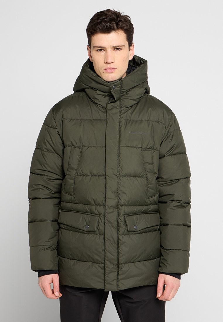 Mann in einer olivgrünen gefütterten Winterjacke mit hohem Kragen und Kapuze, der vor einem schlichten hellen Hintergrund steht.