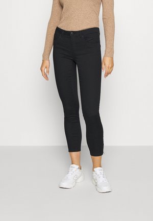 Mujer vestida con vaqueros negros ajustados y recortados, camiseta beige de manga larga y zapatillas blancas, de pie frente a un fondo claro y liso.