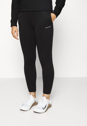 Persoon die zwarte Calvin Klein leggings met zijstrepen en witte Nike sneakers draagt, staand tegen een effen witte achtergrond.