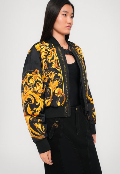 Veste bomber noire avec des motifs floraux dorés, poignets et ourlet côtelés. Présente des détails de marque le long de la fermeture éclair avant et un design court.