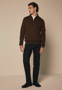 Brauner Strickpullover mit Reißverschlusskragen, kombiniert mit schwarzen Slim-Fit-Hosen und schwarzen Schuhen. Das Model steht vor einem beigen Hintergrund.