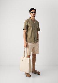 Strikket kortærmet skjorte i olivengrøn, beige shorts, brune sandaler og en cremefarvet totebag med håndtag, stående foran en neutral baggrund.