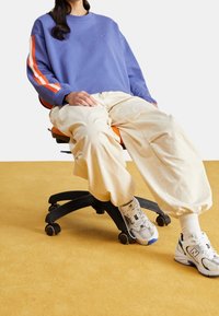 Una persona vestita con una felpa blu e pantaloni beige seduta su una sedia da ufficio arancione con ruote nere su un pavimento giallo.