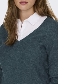 V-Ausschnitt-Pullover aus dunkelblauer Maschenware, kombiniert mit einem weißen Hemd mit Kragen. Weiche Textur und lässiges Design mit einer lockeren Passform.