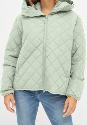 Personne portant une veste matelassée vert clair zippée avec capuche et un jean bleu, debout les mains détendues le long du corps.