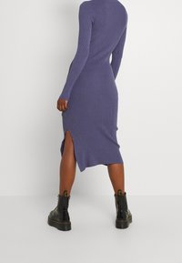 Vestito lungo a coste di colore viola scuro, con una vestibilità aderente, maniche lunghe e uno spacco laterale. Abbinato a stivali neri alla caviglia.