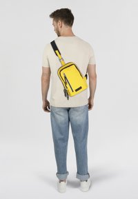 Bolso bandolera de tela amarilla brillante, de forma rectangular con acentos negros, que cuenta con un bolsillo delantero con cremallera y correa ajustable.
