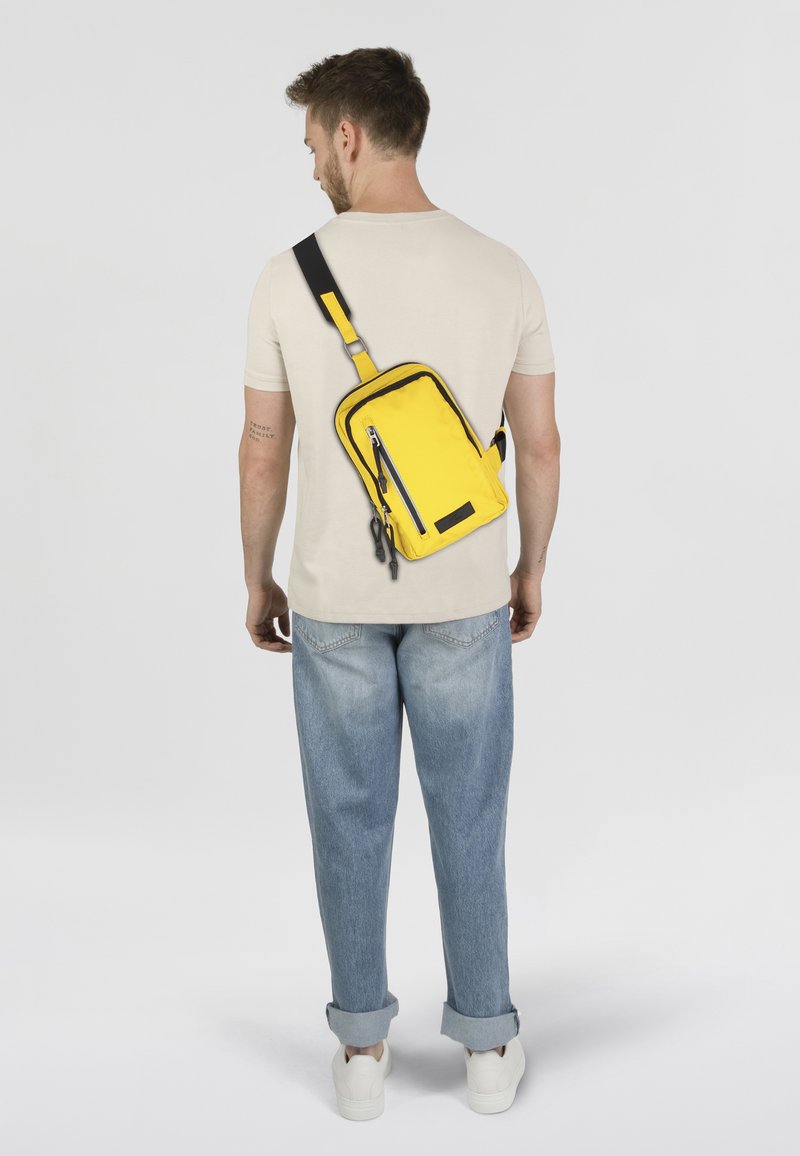 Bolso bandolera de tela amarilla brillante, de forma rectangular con acentos negros, que cuenta con un bolsillo delantero con cremallera y correa ajustable.