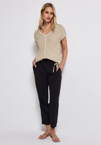 Beige bluse med V-hals, korte ærmer og hvid kant, parret med sorte bukser med snor i livet og flade sandaler. Blødt stof og afslappet pasform.