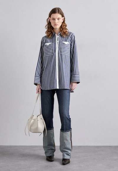 Camicia a righe blu e bianca con due tasche sul petto; abbinata a pantaloni in denim blu e a una borsa a secchiello di colore chiaro.