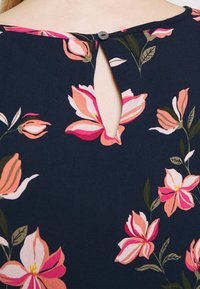 Laivastonsininen paita, jossa on vaaleanpunaisia ja kermaisia kukkakuoseja, avainreikäkaulus ja koristebutton takana. Pehmeä kangas.