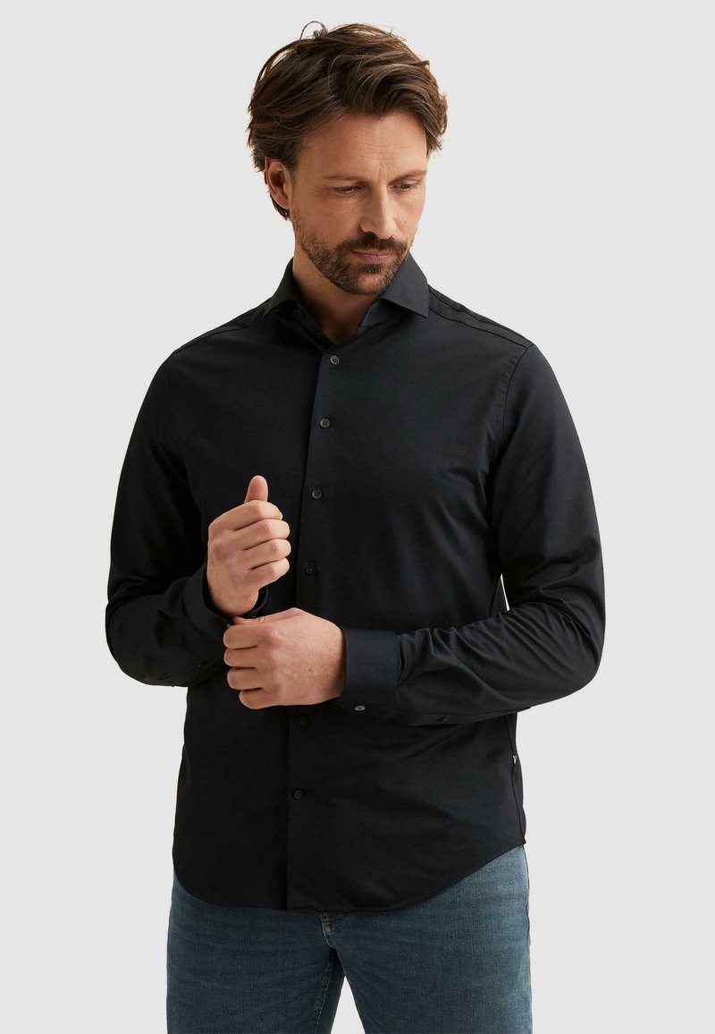 Homme aux cheveux courts châtain et à la barbe ajustant le poignet d'une chemise noire à manches longues boutonnée, portant un jean bleu, fond neutre.