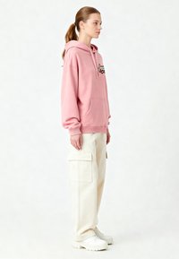 Felpa rosa chiaro con tasca a marsupio e logo, abbinata a pantaloni cargo color crema. Calzature bianche. Tessuto liscio, vestibilità rilassata.