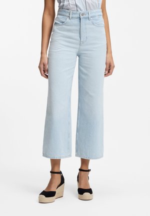 Mujer con vaqueros azules claros de talle alto, pata ancha y tobilleros, y sandalias de cuña tipo alpargata negras con tiras en el tobillo, de pie sobre un fondo blanco.