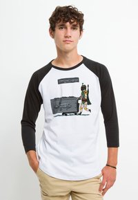 T-shirt bianca da baseball con maniche raglan nere, caratterizzata da una grafica di un personaggio che spinge bagagli sovradimensionati con un design umoristico.