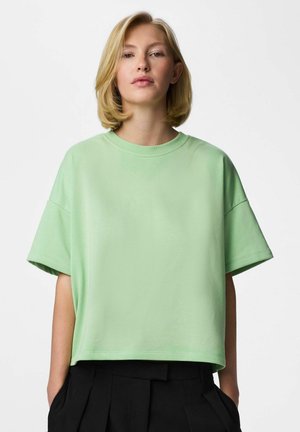 Ung kvinde med kort blondt hår iført en løs, lysegrøn kortærmet skjorte og sorte bukser mod en ensfarvet baggrund.