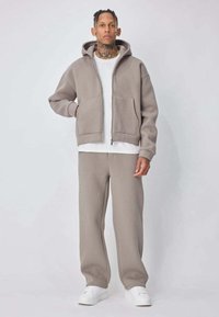 Homme tatoué portant un sweat à capuche zippé gris, un pantalon de survêtement assorti, un T-shirt blanc et des baskets blanches, debout devant un fond uni.