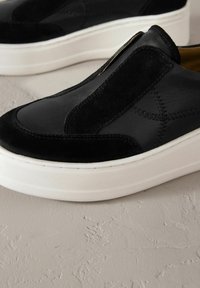 Black slip-on shoes featuring a mix of smooth leather and suede, with a white rubber sole and decorative stitching details.