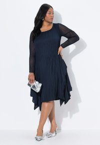 Vestido de color navy con pliegues, mangas largas transparentes, dobladillo asimétrico, tejido texturizado y accesorios plateados que incluyen un bolso de mano y zapatos con lazos.