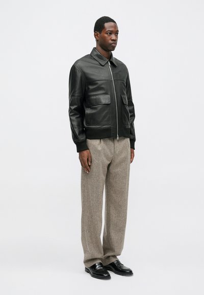 Giacca bomber in pelle nera con due tasche frontali, polsini e colletto a coste, abbinata a pantaloni in tessuto tweed di colore chiaro e scarpe nere.