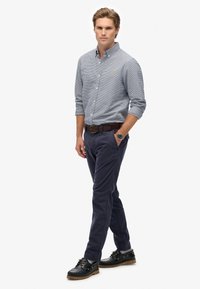 Overhemd met knopen in een blauw geruit patroon, marineblauwe broek, leren bruine riem en zwarte schoenen met gouden accenten. Model staat voor een neutrale achtergrond.