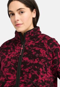 Giacca in pile con un motivo astratto rosa e nero, colletto alto, chiusura con zip e un piccolo logo sul petto. Polsini elasticizzati e tasche.