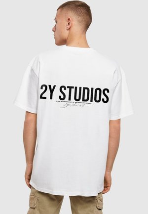 Bílá oversized tričko s černým potiskem na zadní straně, obsahující nápis "2Y STUDIOS" a podpisový detail. Krátké rukávy a kulatý výstřih.
