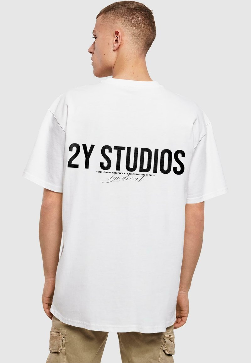 Bílá oversized tričko s černým potiskem na zadní straně, obsahující nápis "2Y STUDIOS" a podpisový detail. Krátké rukávy a kulatý výstřih.