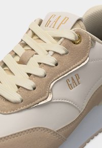 Κοντινή όψη μπέζ και λευκού αθλητικού παπουτσιού με κρεμ κορδόνια και χρυσό λογότυπο "GAP" στη γλώσσα και στην πλαϊνή πλευρά.