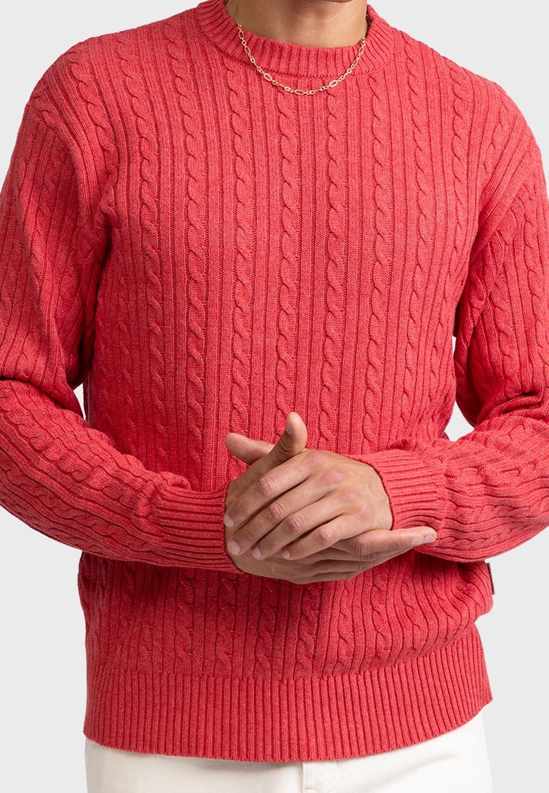 Červený pletený svetr s copánkovým vzorem, žebrovanými manžetami a lemem, kulatým výstřihem. Textura působí měkce a pružně.