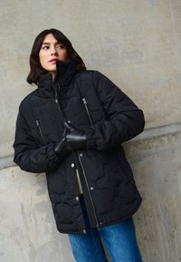 Čierna prešívaná puffa bunda s vysokým golierom, zipsovými vreckami a elastickými manžetami. Nosená s modrými džínsami na pozadí z betónu.