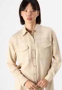Camicia beige con bottoni, dotata di due tasche sul petto, colletto e dettagli di cucitura leggeri. Tessuto liscio, maniche lunghe, realizzata in tessuto leggero.