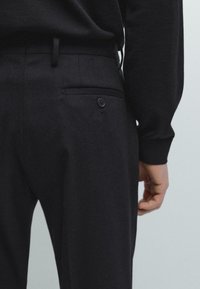 Svarta skräddarsydda byxor med en slät yta, med en bakficka med en svart knapp. Midjan har en minimalistisk design.