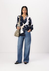 De blouse met lange mouwen en knopen heeft abstracte patronen in marineblauw, crème en lichtblauw, gecombineerd met wijde lichtblauwe denimjeans en een grijze schoudertas.