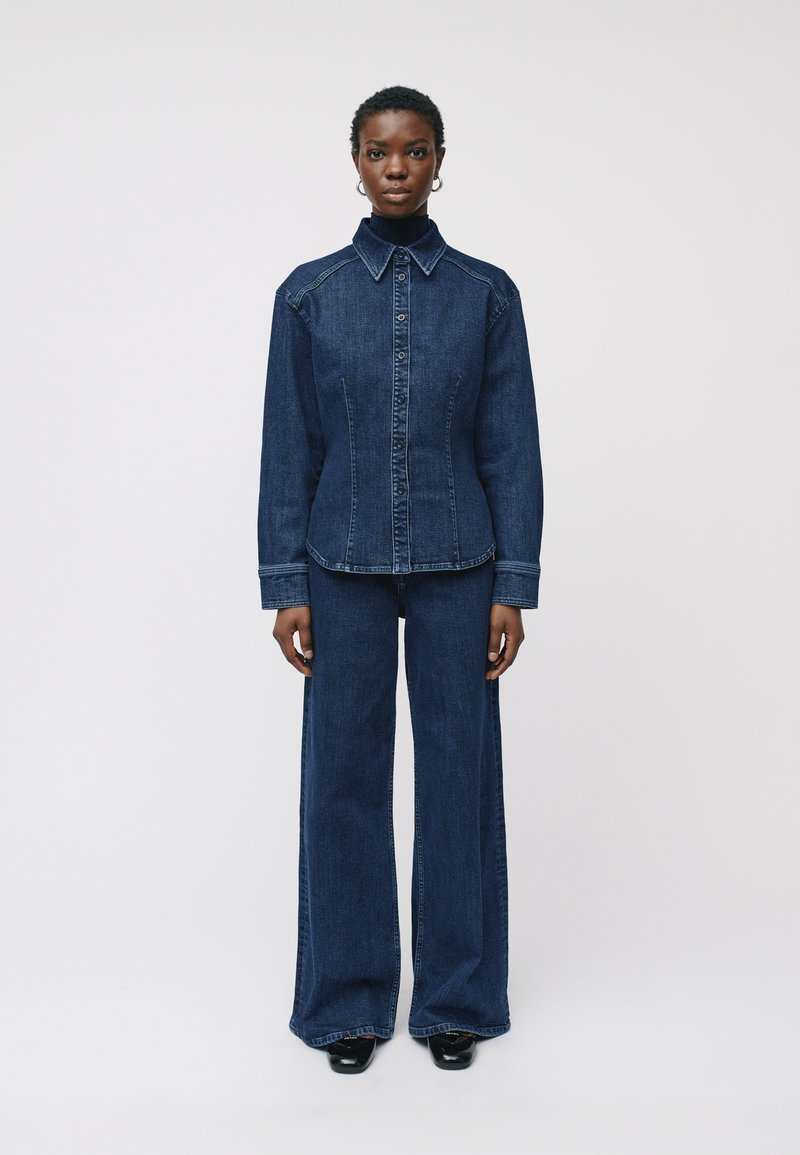 Camicia di denim con bottoni frontali, colletto a punta e vestibilità strutturata. Abbinata a pantaloni in denim a zampa coordinati. Colore blu scuro.