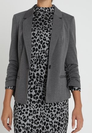 Kvinde iført en tætsiddende grå blazer over en langærmet sort og grå kjole med leopardprint, armene afslappet langs siden.
