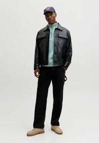 Giacca di pelle nera con due tasche sul petto, maglione verde chiaro con zip, jeans neri e scarpe beige; cappellino blu navy con un logo.