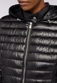 Svart vadderad jacka med glänsande yta, dragkedja och huva med snoddar. Designen inkluderar horisontella sydda paneler.