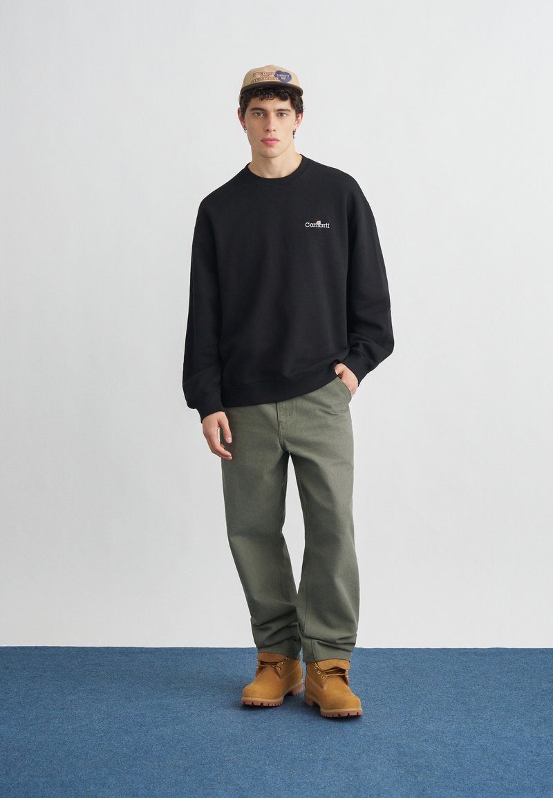 Giovane uomo con cappellino beige, felpa nera Carhartt, pantaloni verdi e stivali da lavoro color sabbia, in piedi su moquette blu con sfondo bianco.